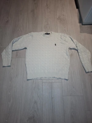 Vit kabelstickad tröja Polo Ralph Lauren - Kabelstickad vit tröja från Polo Ralph Lauren med klassisk rund halsringning och den ikoniska broderade loggan på bröstet. Tröjan har långa ärmar och ribbade muddar vid ärmslut och nederkant. Perfekt för en clean och stilren look.
