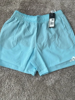 Ljusblå träningsshorts från Adidas - Snygga ljusblå träningsshorts från Adidas med elastisk midja och diskret logga på benet. Shortsen har en enkel och sportig design, perfekta för sommarens alla aktiviteter. Tillverkade i lätt och snabbtorkande syntetmaterial.
