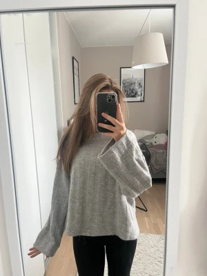 Grå  stickad tröja - En grå stickad tröja med breda ärmar och avslappnad passform. Tröjan har ett diskret ribbmönster och rund halsringning. Perfekt för dig som gillar en chill och trendig look. Materialet ser ut att vara mjukt och bekvämt.