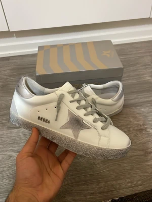Golden Goose skor - Snygga vita sneakers från Golden Goose med grå stjärna på sidan, grå skosnören och silverfärgade detaljer vid hälen. Skorna har en slitstark sula med mönster och är tillverkade i läder. Perfekta för dig som vill ha en trendig och unik look.