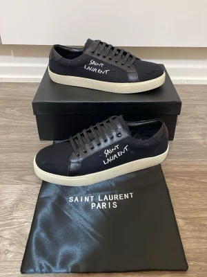 Saint Laurent sneakers - Säljer ett par svarta Saint Laurent sneakers med vit sula och handskriven logga på sidan. Skorna har lågt skaft, rund tå och snörning. Materialet är en mix av skinn och textil som ger en clean och stilren vibe.