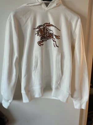 Vit burberry  hoodie med brunt tryck - Säljer en vit hoodie från Burberry med ett stort brunt och svart tryck av en riddare på bröstet. Hoodien har huva med dragsko och en stor magficka framtill. Tillverkad i mjukt bomullsmaterial och har ribbade muddar vid ärmslut och nederkant.