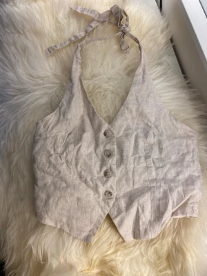 Beige linneväst med knyt och knappar - Snygg beige väst i linne med knyt i nacken och v-ringad front. Västen har fyra knappar framtill och en croppad passform som ger en trendig look. Perfekt att styla över en t-shirt eller på egen hand för en cool sommarstil.