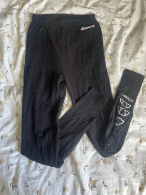 Svarta leggings från Ellesse - Snygga svarta leggings från Ellesse med logga på höften och vita tryck längs ena benet. Stretchigt material som sitter tight och bekvämt. Perfekta till en sportig eller avslappnad look.