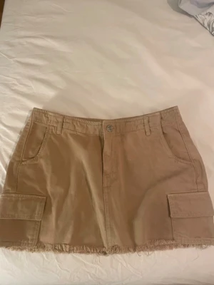 Beige kortkjol med fickor - Säljer en beige kortkjol i bomull med rå kant nertill och stora cargofickor på sidorna. Kjolen har bälteshällor, knapp och dragkedja framtill samt fickor både fram och bak. Perfekt för en avslappnad och trendig look.