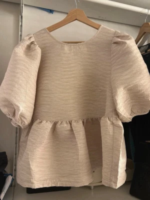 Beige blus med puffärm och knyt - Supersöt beige blus från Carin Wester med puffiga ärmar och volang nedtill. Blusen har en  v-ringning i ryggen med knytband och är helt oanvänd! Orginal priset är 599kr men säljer för 250