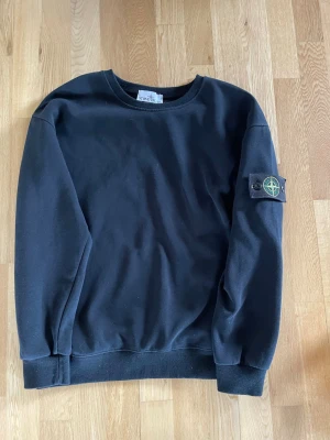 Svart sweatshirt från Stone Island - Säljer en svart sweatshirt från Stone Island med den ikoniska kompass-loggan på vänster ärm. Tröjan har rund halsringning, ribbade muddar och är tillverkad i mjukt bomullsmaterial. Perfekt för dig som gillar streetwear och vill ha en clean look.
