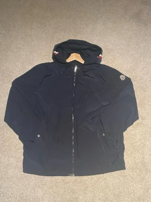 Mörkblå vindjacka från Moncler - Snygg mörkblå vindjacka från Moncler med huva och dragkedja framtill. Jackan har två fickor med tryckknappar och Moncler-logga på vänster ärm. Tillverkad i ett lätt och vindtätt material, perfekt för blåsiga dagar.