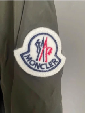 Grå vindjacka från Moncler - Snygg grå vindjacka från Moncler med huva och dragkedja framtill. Jackan har Moncler-logga på ärmen och en cool detalj med röd, vit och blå rand på huvan. Tillverkad i lätt och tåligt material, perfekt för blåsiga dagar.