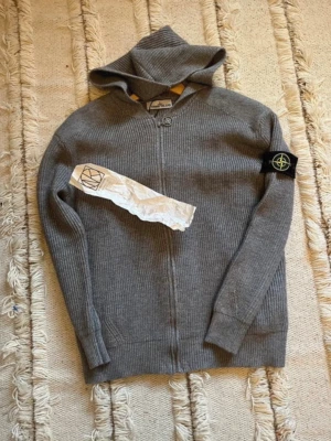 Grå zip hoodie från Stone Island - Säljer en grå stickad hoodie från Stone Island med dragkedja framtill och ribbstickad struktur. Tröjan har huva, långa ärmar och den klassiska Stone Island-loggan på vänster ärm. Perfekt för dig som gillar streetwear och snygga detaljer.