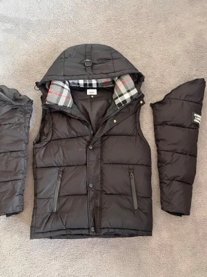 Svart dunväst med avtagbara ärmar - Snygg svart dunväst  med avtagbara ärmar och huva. Insidan av huvan har klassiskt rutigt -mönster i grått, vitt och rött. Två dragkedjeförsedda fickor framtill och quiltad design. Perfekt för lager-på-lager och streetstyle.