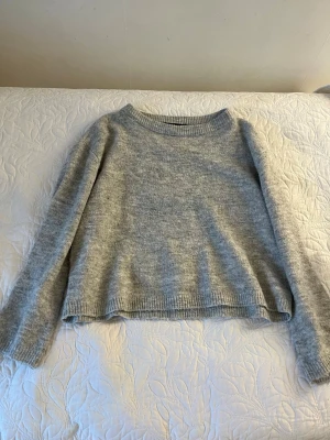 Ljusgrå stickad tröja - Mysig ljusgrå stickad tröja med rund halsringning och långa ärmar. Tröjan har ribbstickade muddar vid ärmslut och nederkant, vilket ger en klassisk och enkel look. Perfekt för lager på lager under kyliga dagar.