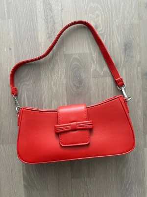 Röd axelväska med dekorativ flik - Cool röd handväska i slät syntet med avtagbar axelrem och dekorativ flik framtill med knutdetalj. Väskan har en stilren, avlång form och stängs med dragkedja. Perfekt för dig som vill ha en färgklick till din outfit.