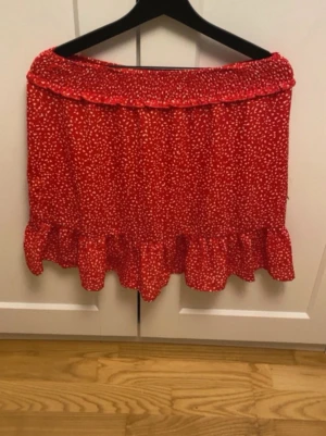 Röd kortkjol med volang och prickar - Supersöt röd kortkjol med vitt prickigt mönster och bred volang längst ner. Kjolen har en smockad midja som ger en bekväm passform och ett lekfullt uttryck. Perfekt för dig som vill ha en färgglad och trendig kjol till sommaren.