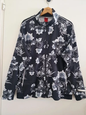 Nike Svart blommig zip-jacka - Nike Snygg svart jacka med hel dragkedja och vitgrått blommönster. Jackan har hög krage och långa ärmar, perfekt för dig som gillar sportig men ändå unik stil. Materialet känns lätt och smidigt, passar till många olika outfits. Använd men fint skick. Storlek 2XL