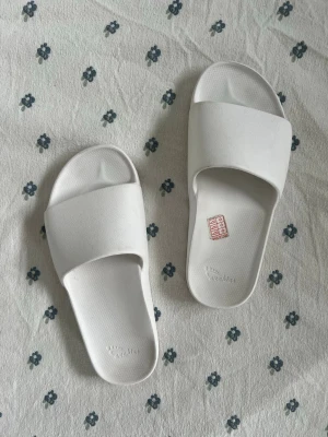 Vita slides från Archies - Säljer ett par stilrena vita slides från Archies. Tofflorna har en bred rem över foten och är gjorda i ett mjukt, flexibelt material som känns bekvämt att ha på sig. Perfekta för att glida runt hemma eller på stranden. Enkel och clean design som passar till allt. Storlek 39.5-40. Har varit med it Vogue. Super trendiga tofflor / sliders. 