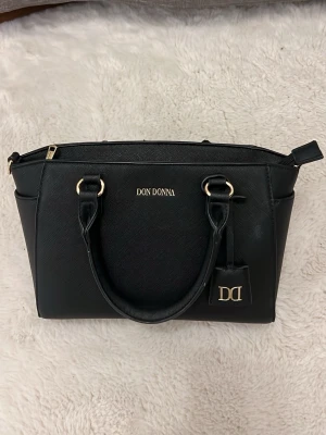 Svart handväska från Don Donna - Stilren svart handväska från Don Donna med guldfärgade detaljer och dubbla handtag. Väskan har en klassisk rektangulär form och är tillverkad i ett tåligt konstläder. Utrustad med dragkedja upptill och en snygg DD-tag som detalj. Finns även band med till väskan. 