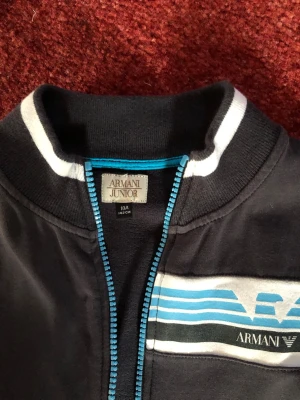 Svart bomberjacka från Armani Junior - Snygg svart bomberjacka från Armani Junior med blå dragkedja och vita detaljer vid krage, ärmslut och nederkant. Jackan har ett tryckt blått och vitt mönster på bröstet och är långärmad. Perfekt för dig som vill ha en stilren och sportig look.använd en gång
