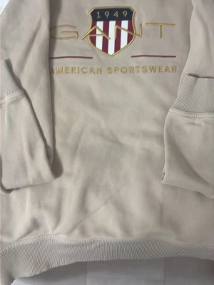 Beige sweatshirt från GANT - Beige sweatshirt från GANT med broderad logga och texten 'American Sportswear' framtill. Tröjan har ribbade muddar vid ärmslut och nederkant samt ett klassiskt collegeinspirerat tryck. Perfekt för en sportig och avslappnad look.
