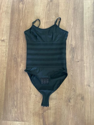 Svart shaping-body med hyskor - Säljer en svart shaping-body med smala axelband och breda förstärkta partier runt midjan för extra stöd. Bodyn har hyskknäppning i grenen och är tillverkad i 90% polyester och 10% elastan för stretch och komfort.