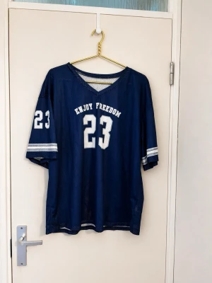 Blå mesh topp med tryck och nummer - Cool blå mesh topp med vitt tryck 'ENJOY FREEDOM' och siffran 23 på bröstet och ärmarna. T-shirten har v-ringning, korta ärmar och vita ränder på ärmsluten. Perfekt för dig som gillar sportig stil och vill sticka ut.