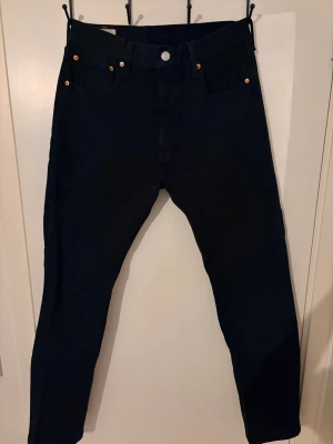 Mörkblå Levi's jeans med smal passform - Säljer ett par mörkblå Levi's jeans med klassisk femficksdesign och smal passform. Jeansen har silverfärgad knapp, nitar och Levi's ikoniska läderpatch bak i midjan. Tillverkade i slitstark denim som passar perfekt till din vardagsstil.