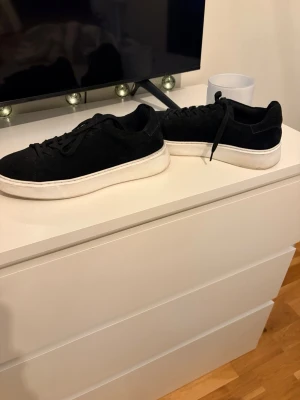 Svarta sneakers med vit sula - Stilrena svarta sneakers med tjock vit sula och svarta skosnören. Ovandelen är i mockaliknande material och designen är klassiskt låg. Perfekta för dig som gillar enkel och clean stil.