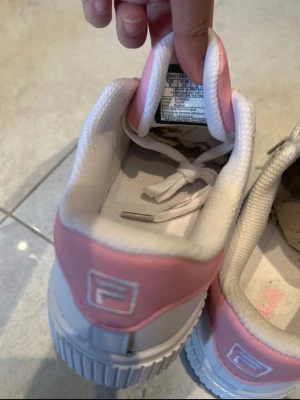 Vita och rosa Fila sneakers strl 35 - Snygga sneakers från Fila i vitt med rosa detaljer runt hälen och på plösen. Skorna har en chunky sula och klassisk låg modell med vita skosnören. Perfekta för dig som gillar sportig och trendig stil.