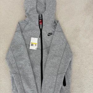 Grå Nike tech  - Snygg grå hoodie från Nike med dragkedja framtill och svart Nike-logga på bröstet. Hoodien har huva, svarta detaljer vid fickorna och är tillverkad i mjukt sweatshirtmaterial. Perfekt för en sportig och avslappnad stil.