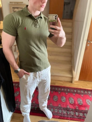 Khakifärgad piké från Ralph Lauren - Säljer en khakifärgad pikétröja från Ralph Lauren med klassisk krage och knappar framtill. Tröjan har korta ärmar och den ikoniska röda logotypen broderad på bröstet. Modellen är custom fit och passar perfekt till en avslappnad men stilren look.