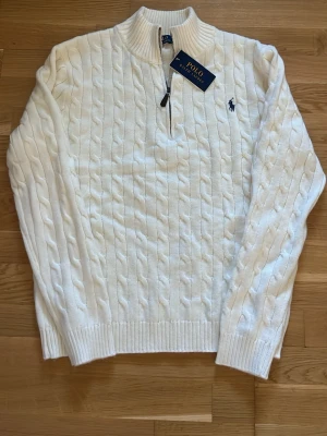Vit kabelstickad tröja Polo Ralph Lauren - Kabelstickad vit tröja från Polo Ralph Lauren med halv dragkedja och ribbad krage. Tröjan har det klassiska blå broderade logotypen på bröstet och ribbade muddar vid ärmslut och nederkant. Perfekt för en stilren och klassisk look.