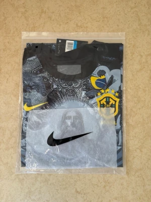 Brazil Nike Pre-Match Jersey (Cristo Redentor / Rio edition) - Snygg t-shirt från Nike med Brasil-logga och stort grafiskt tryck av Kristusstatyn och detaljer i svart, grått och gult. Kortärmad modell med rund hals och coola mönster över hela tröjan. Perfekt för dig som gillar fotboll och unik design.