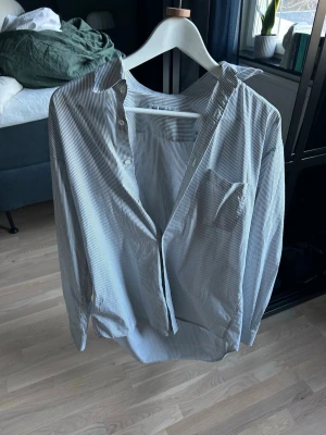 Randig skjorta från Balenciaga - Säljer en stilren vit skjorta med tunna svarta ränder från Balenciaga. Skjortan har klassisk krage, bröstficka och knappar framtill. Perfekt oversized fit för en avslappnad look. Tillverkad i mjuk bomull.