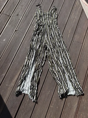 Randig jumpsuit med vida ben - Säljer en snygg jumpsuit med svarta och vita vertikala ränder. Modellen har smala axelband och vida ben för en avslappnad look. Materialet känns lätt och luftigt, perfekt för varma dagar. Inget synligt märke på plagget.