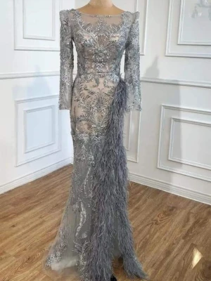 Silverfärgad långklänning med fjädrar - Elegant långklänning i silver med broderade detaljer och transparenta partier. Klänningen har långa ärmar, vacker spets och dekorativa fjädrar längs sidan. Perfekt för dig som vill sticka ut på bal eller fest.