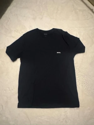 Svart t-shirt från BOSS - En stilren svart t-shirt från BOSS med diskret logga på bröstet. Klassisk passform och rund halsringning, perfekt till jeans eller shorts. Tillverkad i mjuk bomull för skön känsla hela dagen.