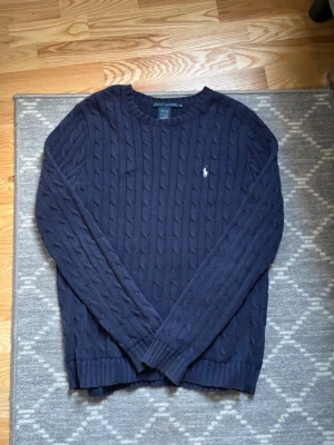 Mörkblå kabelstickad tröja Polo Ralph Lauren - Mörkblå kabelstickad tröja från Polo Ralph Lauren med klassisk rund halsringning och diskret broderad logotyp på bröstet. Tröjan har långa ärmar och ribbade muddar vid ärmslut och nederkant. Perfekt för dig som gillar stilren och tidlös design.