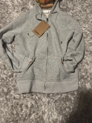 Burberry  hoodie - Snygg grå hoodie från Burberry med dragkedja framtill och klassiskt rutigt foder i huvan. Hoodien har fickor på sidorna, ribbade muddar och justerbar dragsko i huvan. Tillverkad i mjukt bomullsmaterial för en skön känsla.