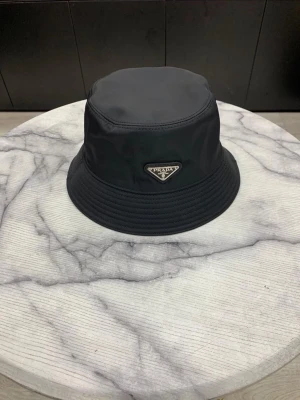 Svart bucket hat - Stilren svart bucket hat i glansigt nylonmaterial. Klassisk form med bred, nedåtlutande brätte och ikonisk metallogo framtill. Perfekt accessoar för en trendig streetstyle-look.