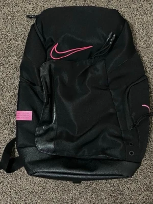 Svart ryggsäck från Nike med rosa detaljer - Snygg svart ryggsäck från Nike med rosa logga och detaljer. Ryggsäcken har flera fack och dragkedjor, samt justerbara axelband. Perfekt för skolan eller träningen och rymmer allt du behöver. Materialet är slitstarkt och vattenavvisande.