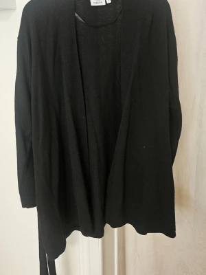 Svart kofta från KappAhl - En svart, långärmad kofta från KappAhl med öppen front och tillhörande knytskärp i midjan. Koftan har en avslappnad passform och är tillverkad i ett mjukt, stickat material som passar perfekt för lager-på-lager-stil.