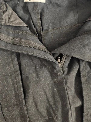 Svarta kostymbyxor med bälte - Säljer ett par svarta kostymbyxor med tillhörande bälte i samma tyg. Byxorna har dragkedja och knappstängning framtill samt pressveck längs benen. Materialet ser ut att vara en blandning av bomull och polyester, vilket ger en klassisk och stilren look.