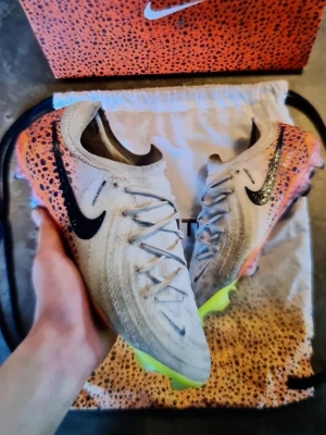Nike Phantom GX Elite fotbollsskor - Säljer ett par Nike Phantom GX Elite fotbollsskor i beige med svarta detaljer och neonfärgade sulor. Skorna har ett coolt leopardmönster i orange och svart på hälen och sidan. Lätta och smidiga med snörning och låg profil, perfekta för snabba vändningar på planen.