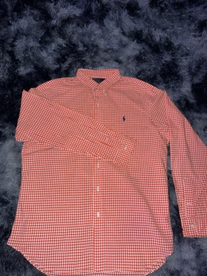 Rutig orange skjorta från Ralph Lauren - Säljer en klassisk orange och vit rutig skjorta från Ralph Lauren med lång ärm och knappar framtill. Skjortan har button-down krage och den ikoniska blå loggan broderad på bröstet. Perfekt för dig som gillar stilrena och tidlösa plagg.