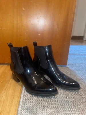 Svarta lackade Chelsea boots - Säljer ett par svarta Chelsea boots i glansigt lackmaterial med elastiska resårpaneler på sidorna och låg klack. Klassisk siluett med dragflikar både fram och bak för enkel påtagning. Perfekta för dig som vill ha en stilren och trendig look.