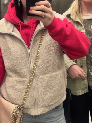 Beige teddyväst med dragkedja - Mysig beige teddyväst med dragkedja framtill och två stora fickor. Västen har en avslappnad passform och är perfekt att slänga över en hoodie för en chill look. Materialet är fluffigt och ger en trendig vibe.