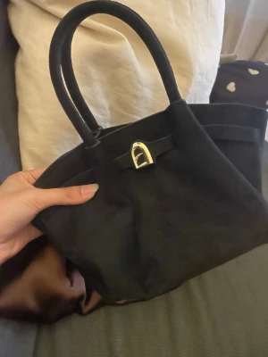 Svart  handväska med scarf - Stilren svart handväska i mocka med två handtag och guldfärgat spänne framtill. Väskan har en brun, glansig scarf knuten runt ena handtaget för en extra touch. Perfekt för dig som gillar klassiska accessoarer med en twist.