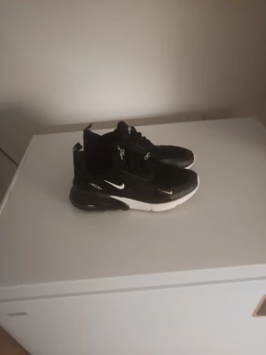 Nike Air Max 270  - Svarta Nike Air Max 270 sneakers med vit sula och röd detalj under sulan. Skorna har tydlig Nike-logga på sidan och textdetaljer. Ovandelen är i meshmaterial för bra andningsförmåga och komfort.