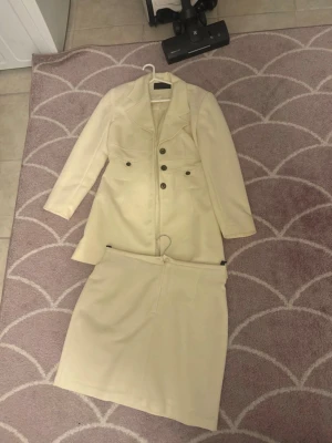 Beige kostym med kavaj och kjol - Stilren beige kostym med långärmad kavaj och matchande kjol. Kavajen har breda slag, dekorativa knappar framtill och markerad midja. Kjolen är rak och enkel i modellen. Perfekt för en clean och klassisk look. Bra skick och jag tycker de är en storlek M/L