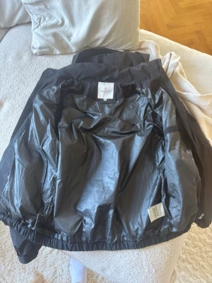 Svart Moncler vindjacka med logotyp - Snygg svart vindjacka från Moncler med dragkedja framtill, huva och två fickor med dragkedja. Det står storlek M men jag tycker passformen är som S! Jackan har en minimalistisk design och elastisk nederkant för skön passform. Perfekt för blåsiga dagar och enkel att matcha med olika outfits. 